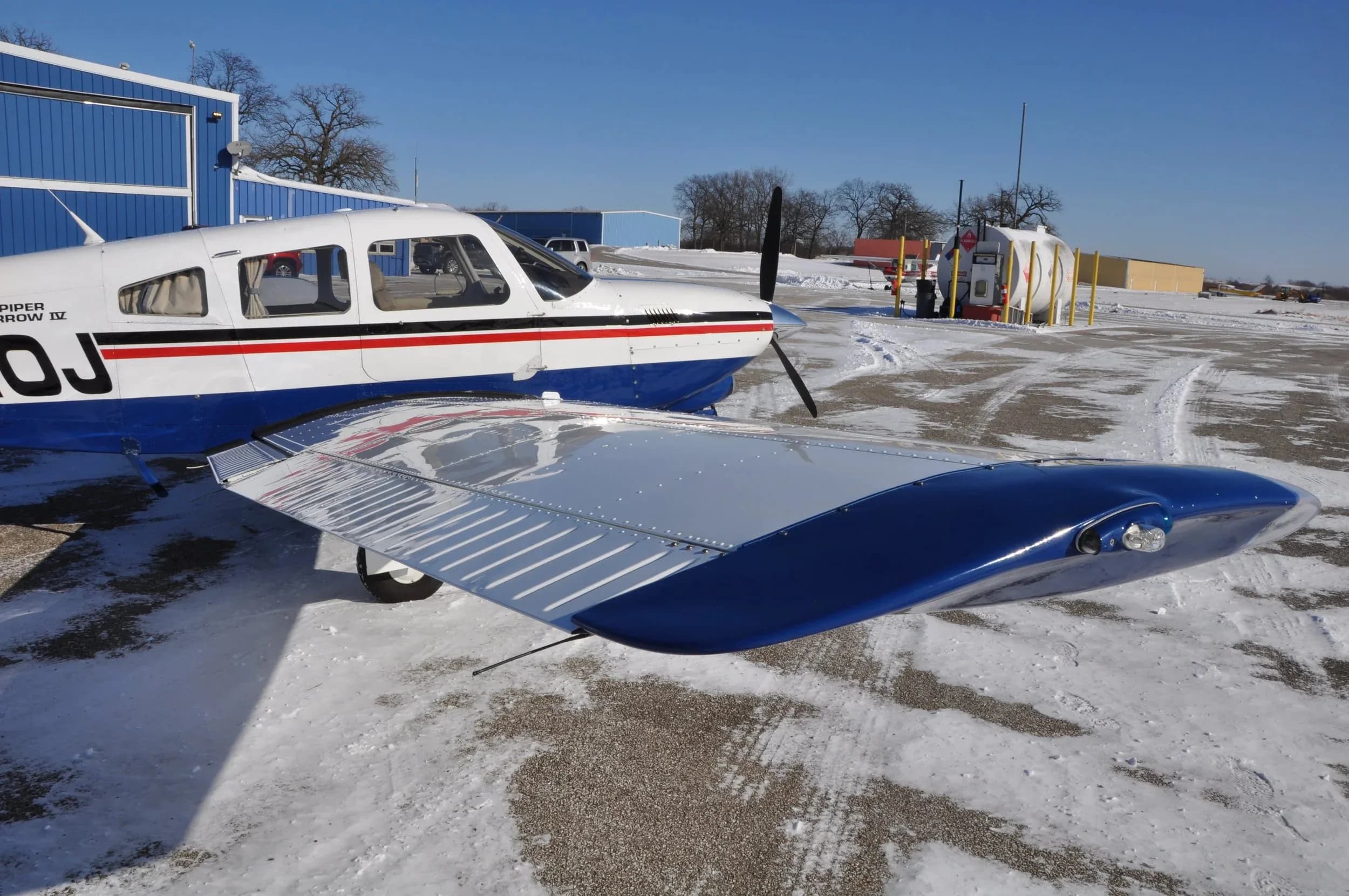 A close-up view of the left wing and nose of a white and blue Piper Arrow IV aircraft with registration N4310J on a snowy tarmac.