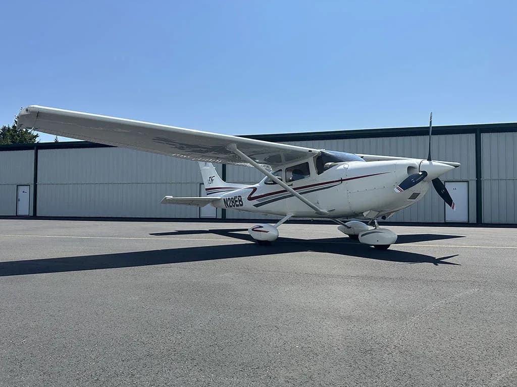 A white Cessna 182 Skylane with red and gray stripes, registration N28EB, parked on an asphalt ramp in front of a hangar.