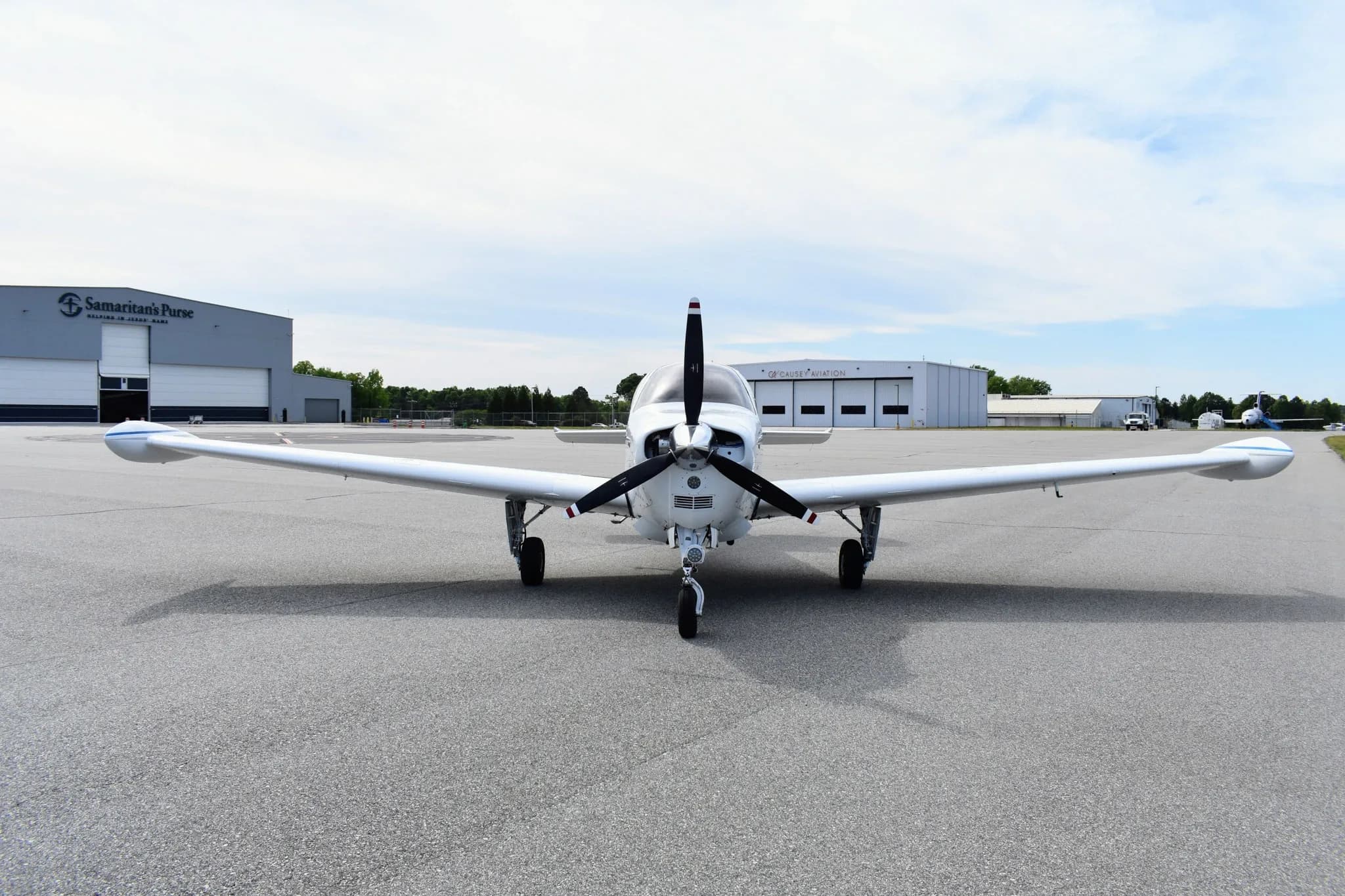 A front view of a white Beechcraft Bonanza F33A with blue and maroon stripes, parked on an asphalt tarmac in front of hangars.