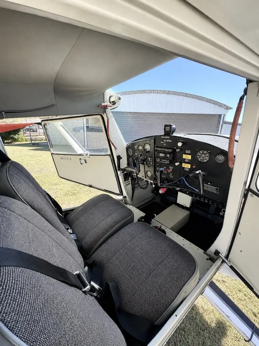 The interior of a small aircraft showing two grey fabric seats and the instrument panel through an open door.