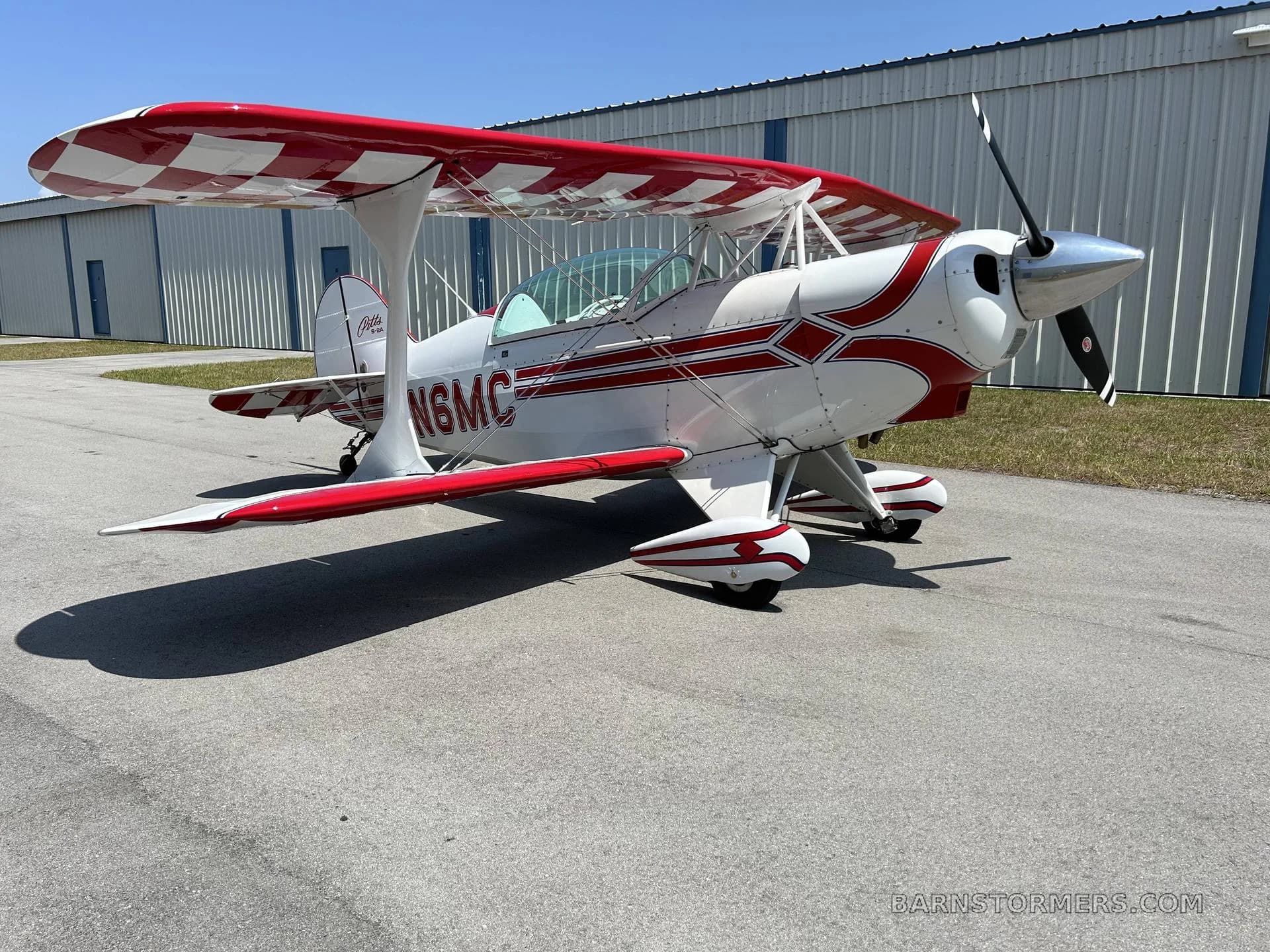 A white Pitts Special biplane with red checkered wings and red stripe accents, parked on an asphalt ramp next to a hangar.