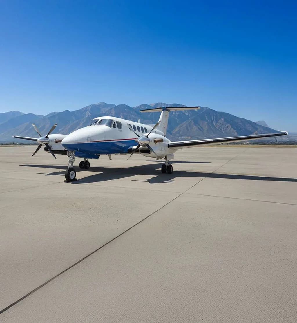 A white and blue turboprop aircraft, likely a King Air, seen from the front on an asphalt runway with mountains in the background.