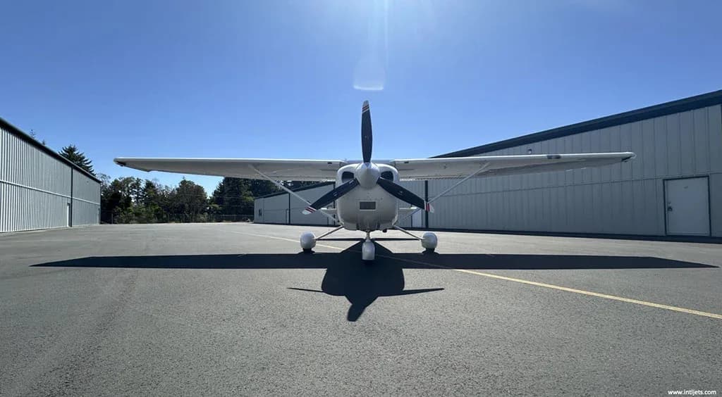 A white Cessna 182 Skylane with red and gray stripes, viewed head-on, parked on an asphalt ramp under a bright sky.