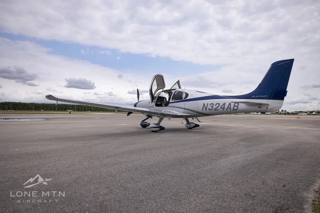 A white and blue Cirrus SR22T aircraft with open doors parked on an airfield, viewed from the rear-left.