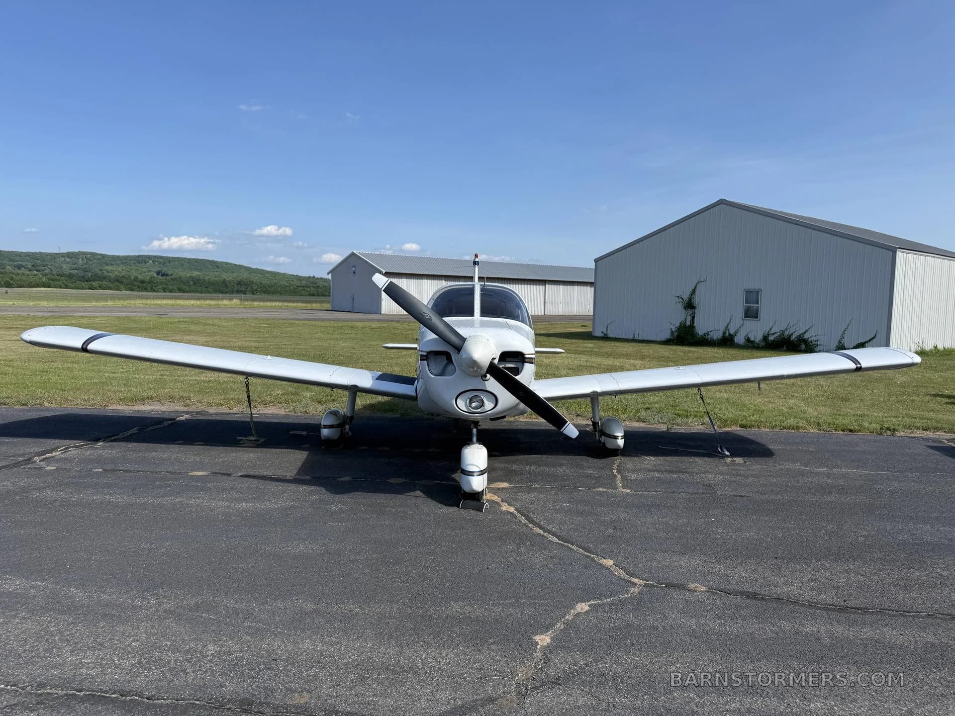 A white single-engine propeller aircraft (N95266) with blue stripes, viewed head-on, parked on an airport apron with hangars behind it.