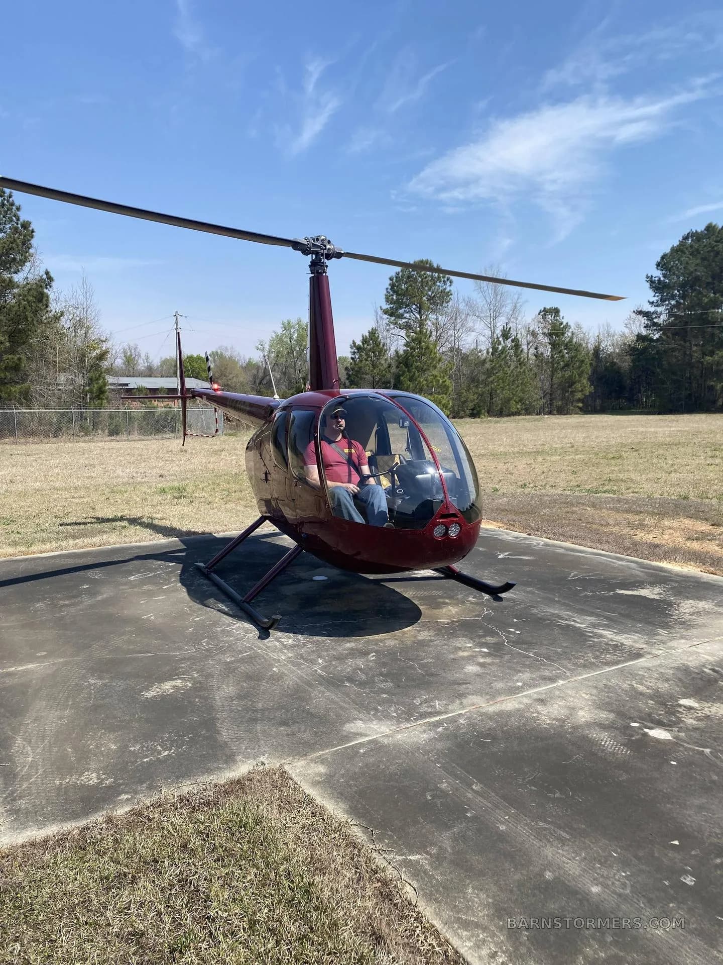 A dark red Robinson R44 Raven II helicopter with a pilot inside, parked on a concrete pad at an airfield under a blue sky.