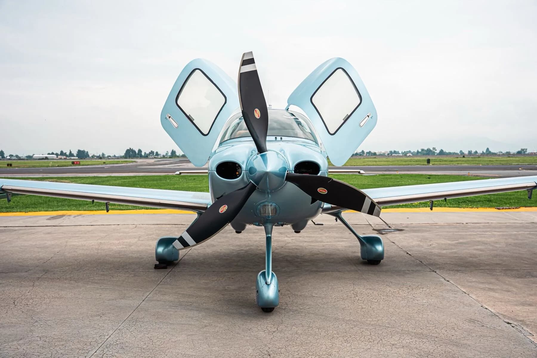 Front view of a light blue Cirrus SR22T aircraft with its gull-wing doors open on an airfield.