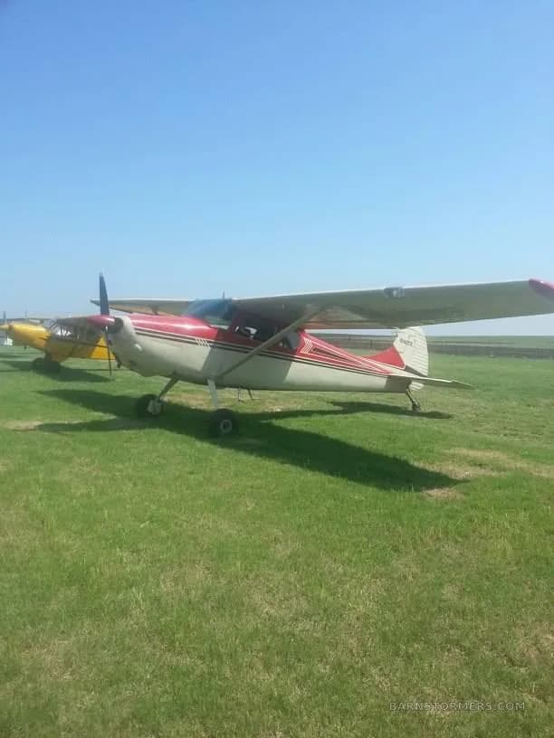 A red and white high-wing single-engine propeller aircraft parked on a grassy field under a clear blue sky, with another yellow aircraft partially visible.