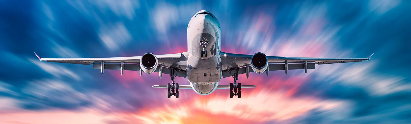 Commercial airliner flying with landing gear down against a colorful sky.
