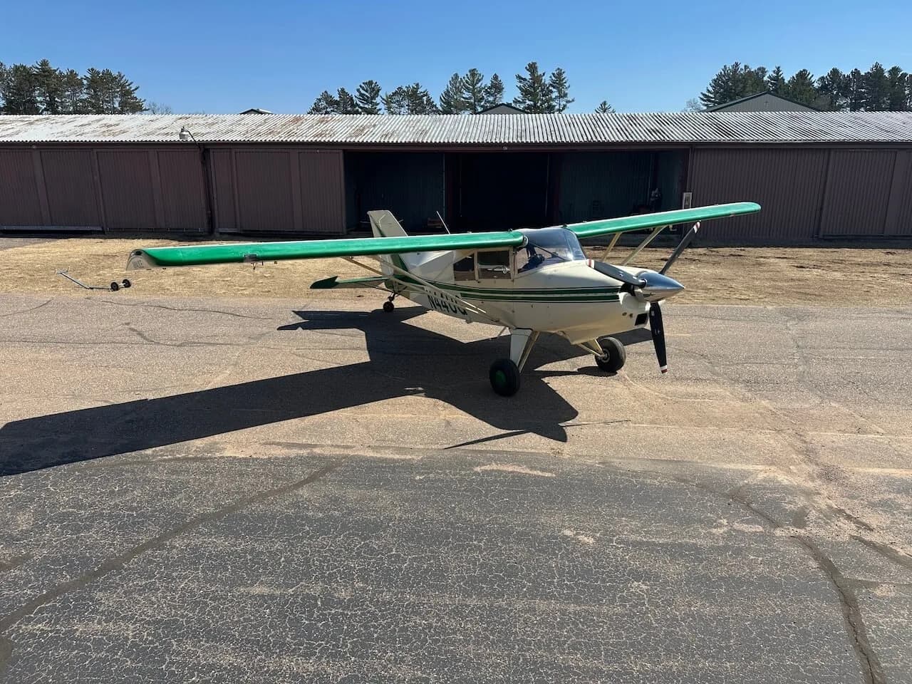 A Maule aircraft with a green and white paint scheme is parked on an asphalt airfield in front of hangars.