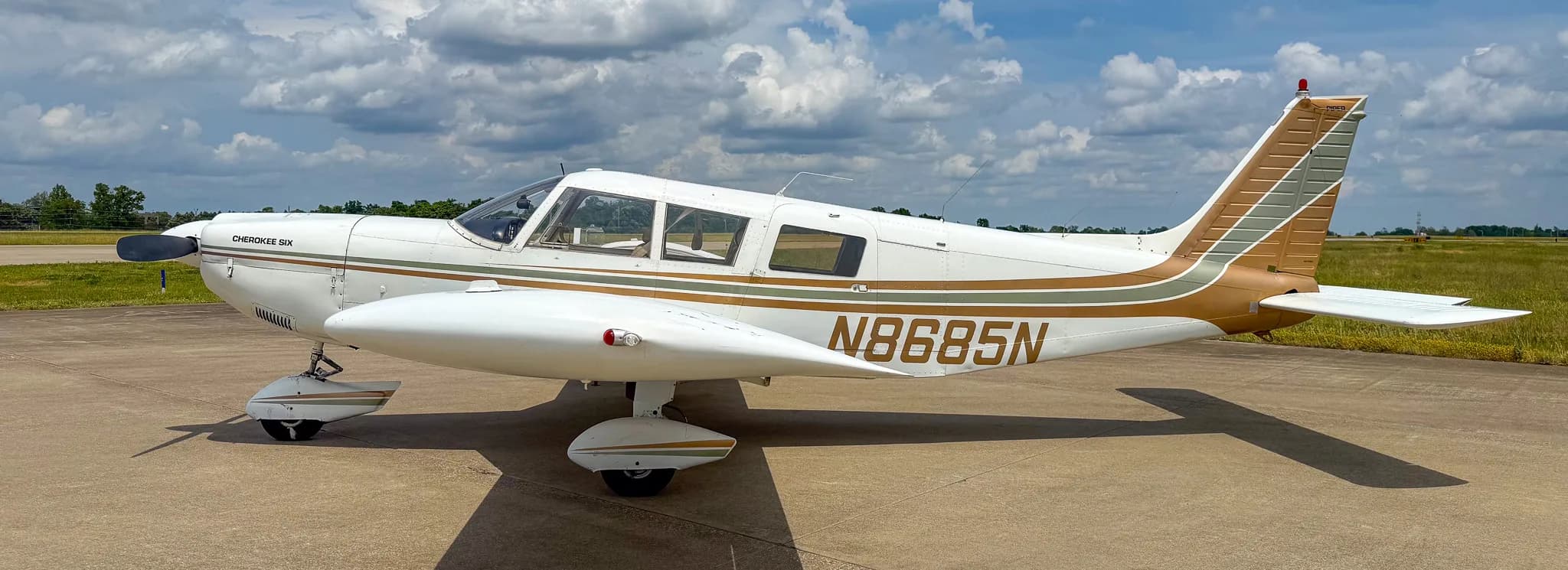 A side view of a white Piper Cherokee Six aircraft with brown and green stripes, showing registration N8685N, parked on an airfield.