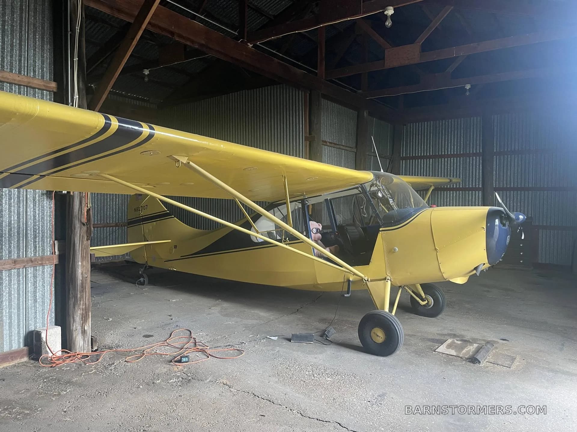 A side view of a yellow Aeronca Champ aircraft with black stripes and a blue cowl, parked in a rustic hangar.