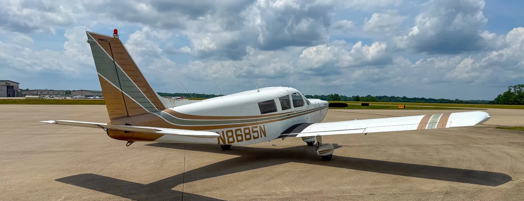 A rear-right view of a white Piper Cherokee Six aircraft with brown and green stripes, showing registration N8685N, parked on an airfield.