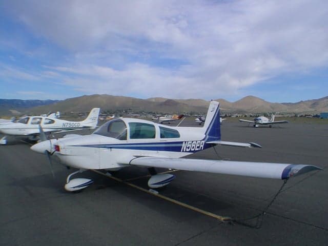 A white and blue Grumman AA-5 Traveler aircraft (N56ER) parked on an airfield with other planes and mountains in the background.