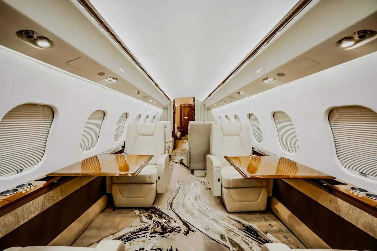 A wide view of a private jet cabin interior with light-colored seats, wooden tables, and a distinctive patterned carpet.