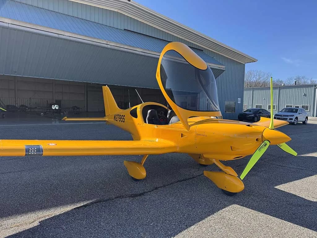 A yellow Bristell aircraft, N279SS, with its canopy open, is parked on the tarmac in front of hangars.