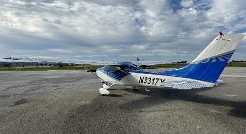A Cessna 182 Skylane aircraft with a blue and white paint scheme is parked on the tarmac at an airport.