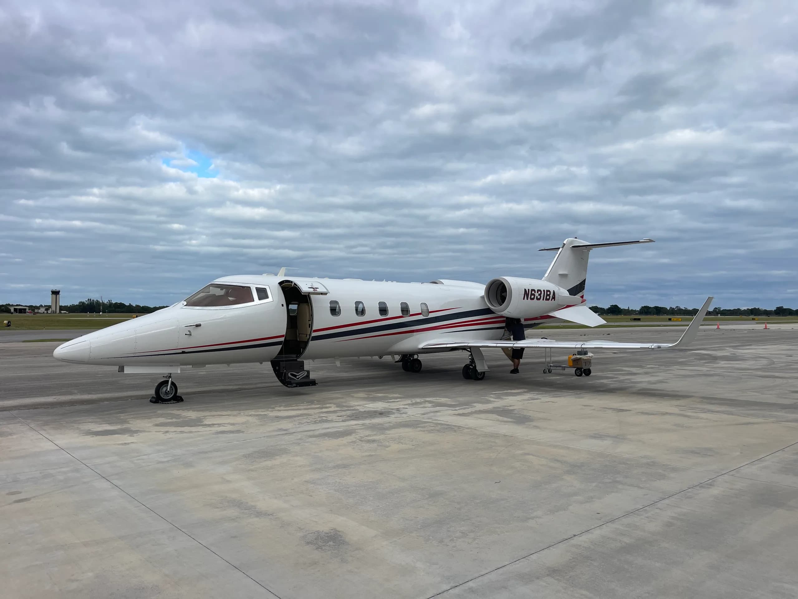 A white Learjet 45/75 with red and black stripes and its door open sits on an airport tarmac under a cloudy sky.