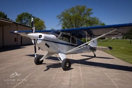 A white high-wing taildragger aircraft with blue accents, registration N620NY, parked on an airfield ramp.