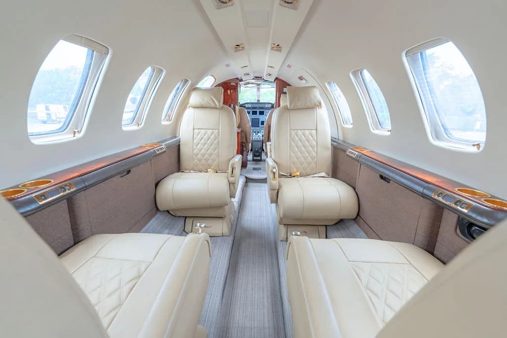 The interior cabin of a private jet with light beige leather seats and wood trim, looking towards the cockpit.