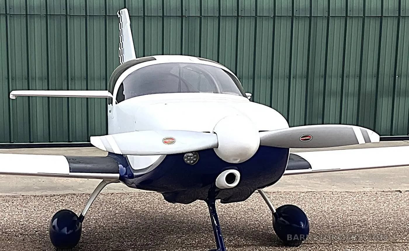 A head-on view of a white and blue single-engine aircraft with a propeller, parked in front of a green hangar.