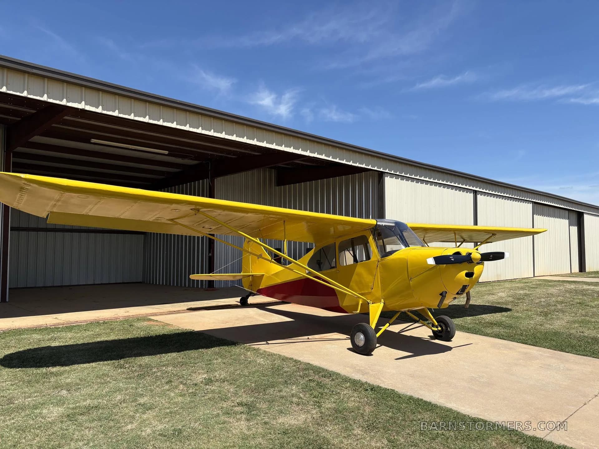 A yellow single-engine aircraft with red accents, parked in front of a hangar under a blue sky.