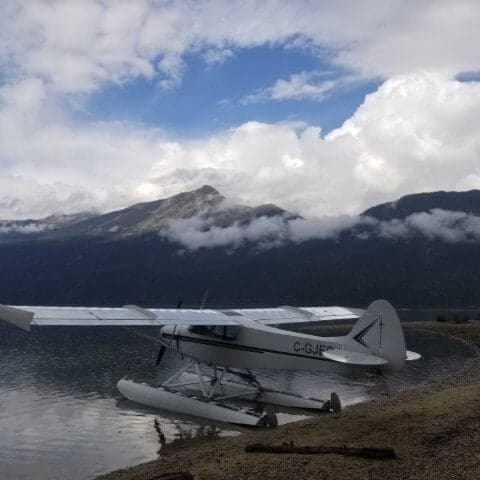 A white and blue Super Cub floatplane on the shore of a lake with mountains in the background.