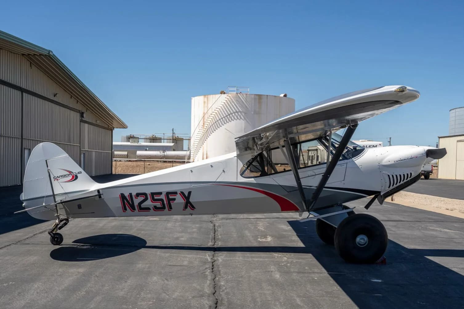 A side view of the white and grey Carbon Cub FX-3 aircraft N25FX parked on an asphalt ramp with large storage tanks in the background.