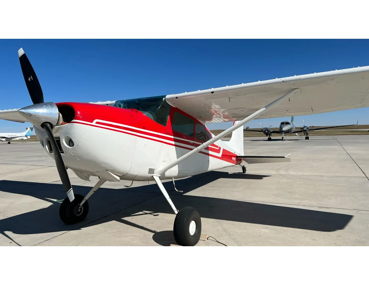 Front-left view of a white and red Cessna 172 on an airport tarmac under a clear blue sky.
