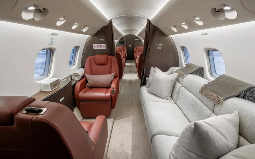 A luxurious private jet cabin featuring a grey sofa on one side and red leather seats on the other, separated by an aisle.