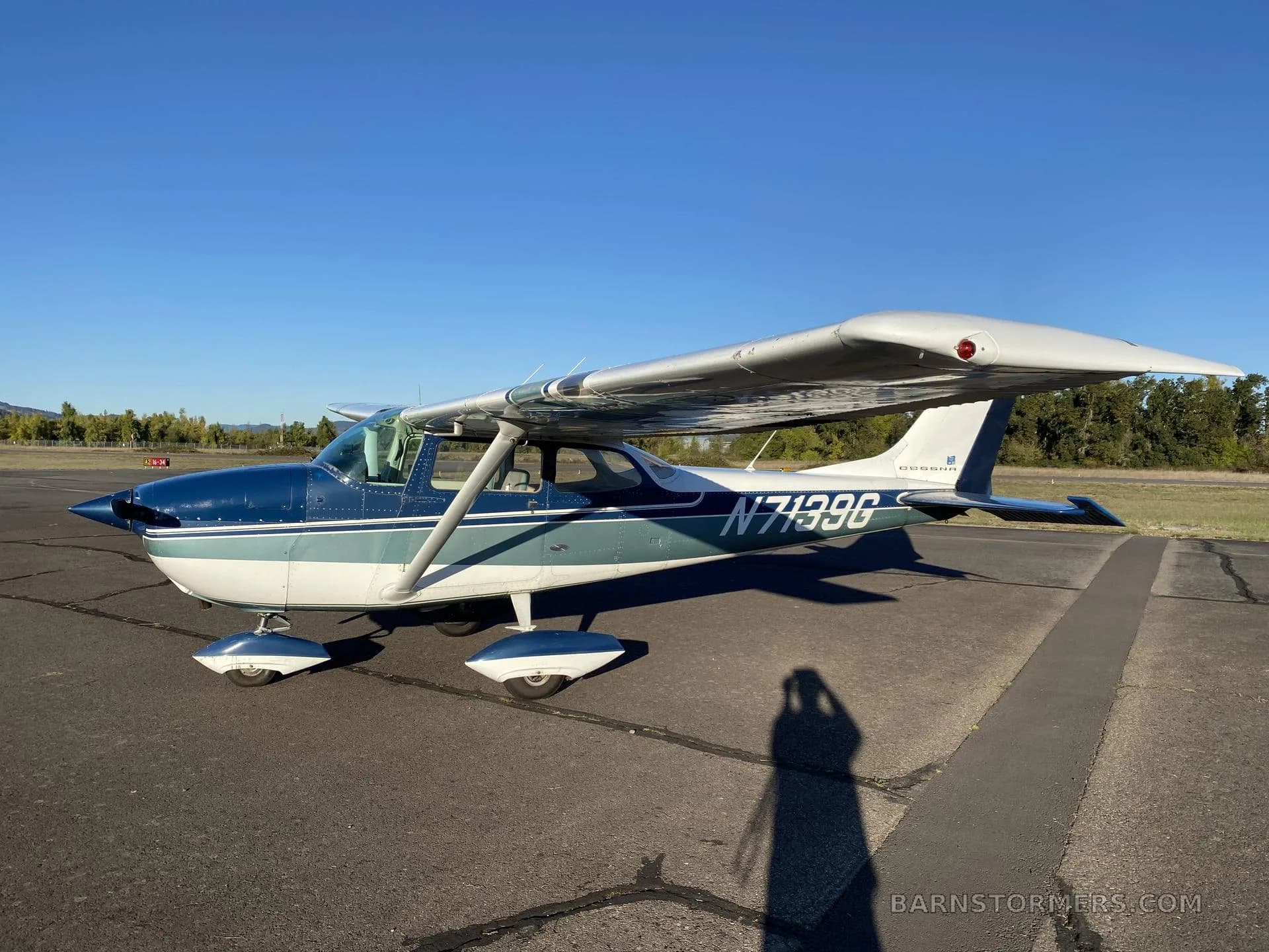 A blue and white Cessna N7199G aircraft is parked on an asphalt ramp, viewed from the side, under a clear blue sky.