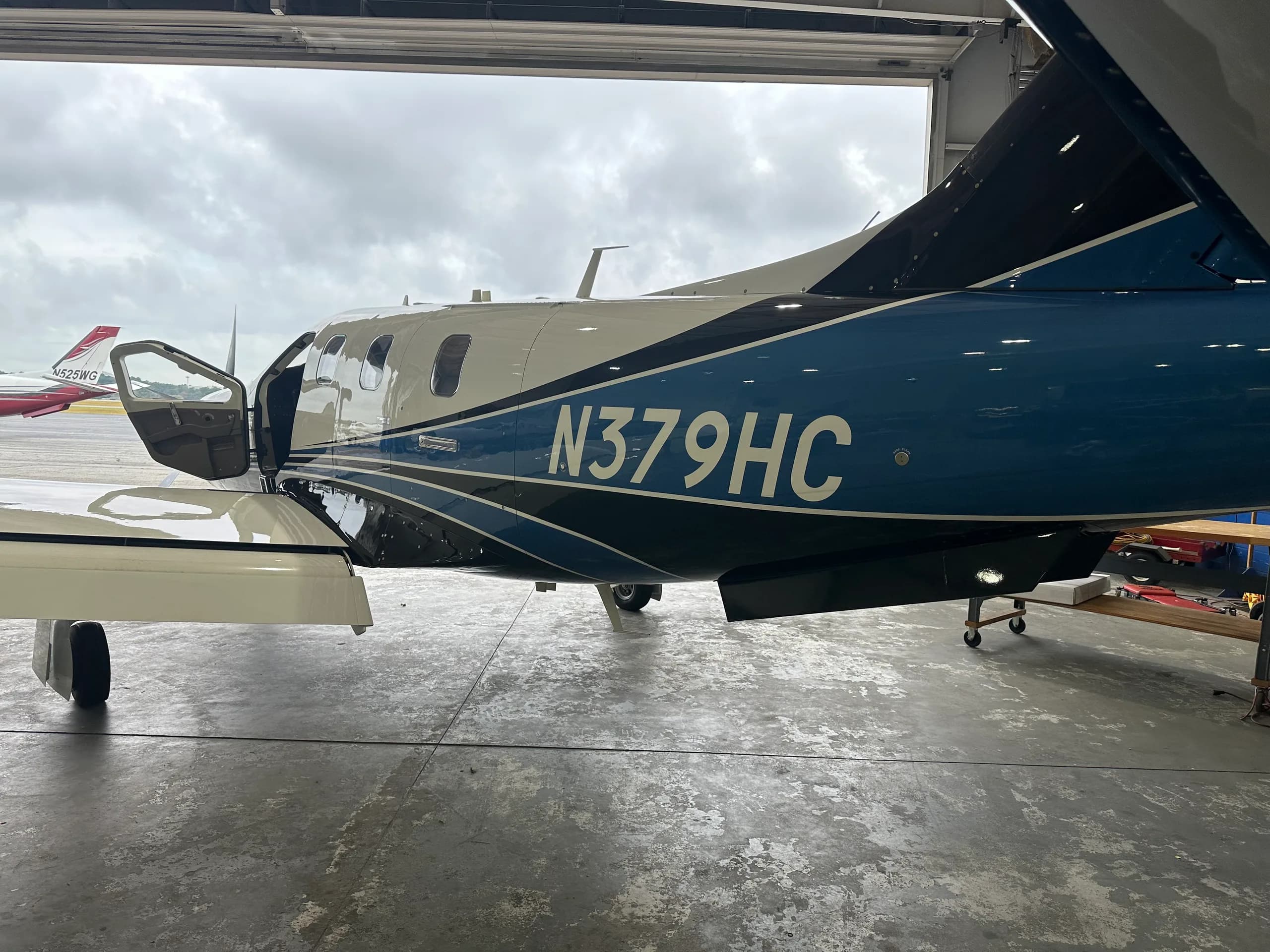 The side of a white and blue TBM 960 aircraft with registration N379HC, parked inside a hangar with its door open.