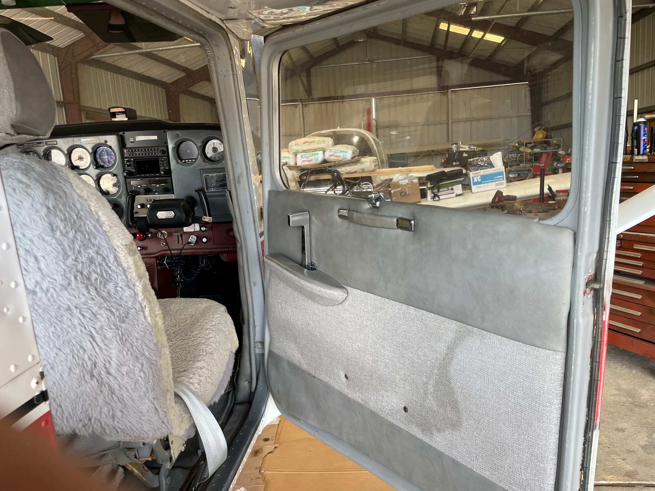 The open door and front cabin area of a Cessna 172, showing a sheepskin-covered seat and part of the instrument panel.