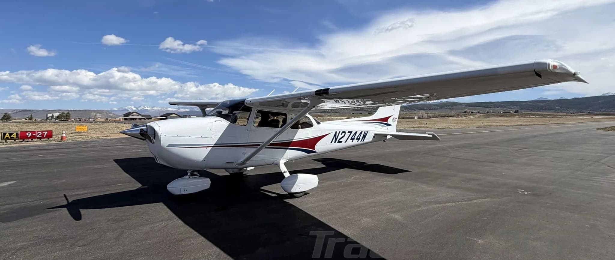 A front-side view of a white Cessna 172 Skyhawk with red and blue stripes, registration N2744W, on an airfield.