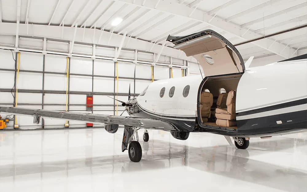 A black and white Pilatus PC-12 aircraft with its main cabin door open, revealing the interior, parked in a hangar.