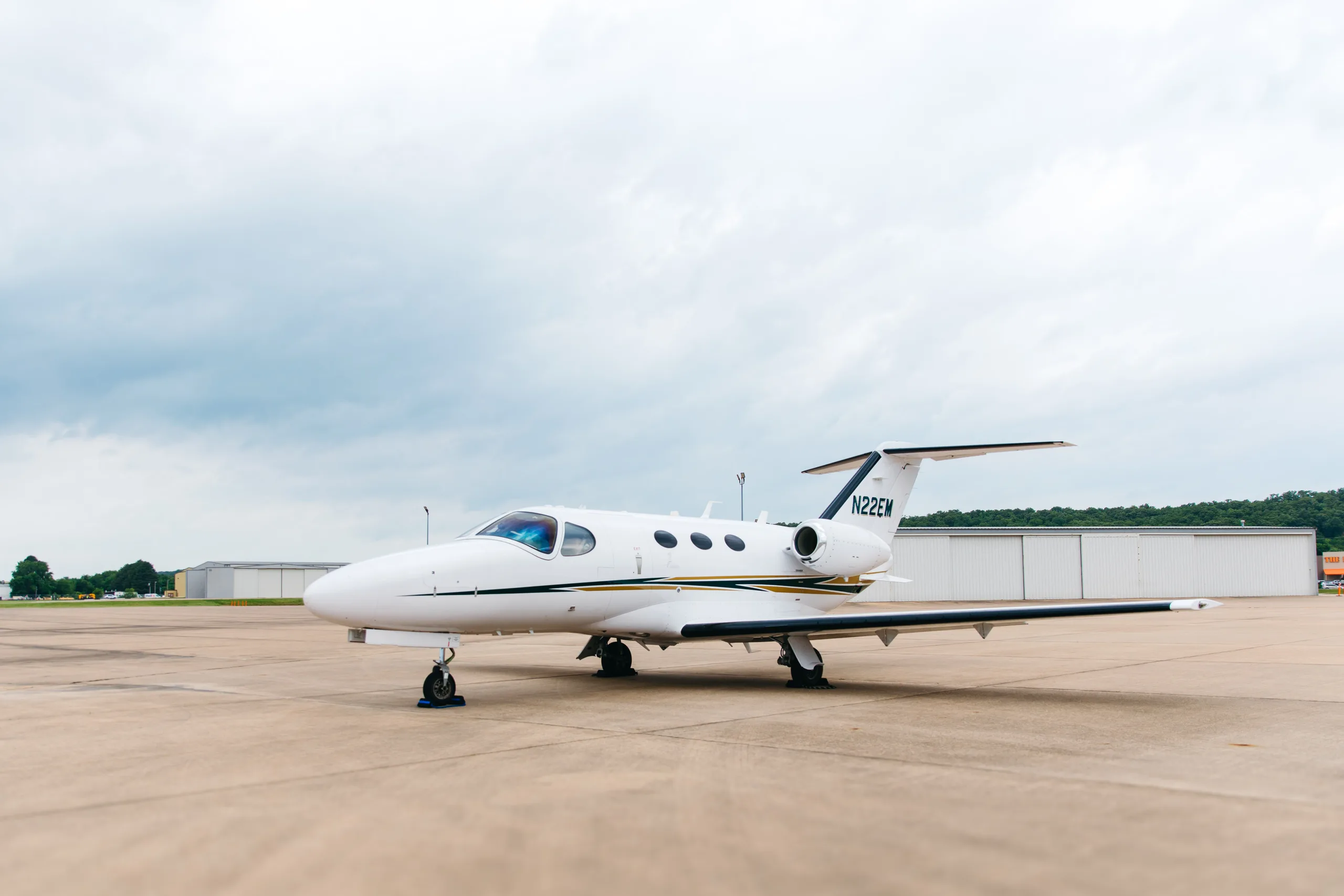 A white Cessna Citation M2 business jet, tail number N22EM, with gold and dark green stripes, parked on a tarmac with hangars in the background.