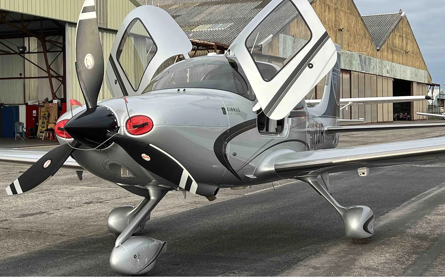 A silver Cirrus SR22 aircraft with black accents and open doors is parked in front of a hangar.