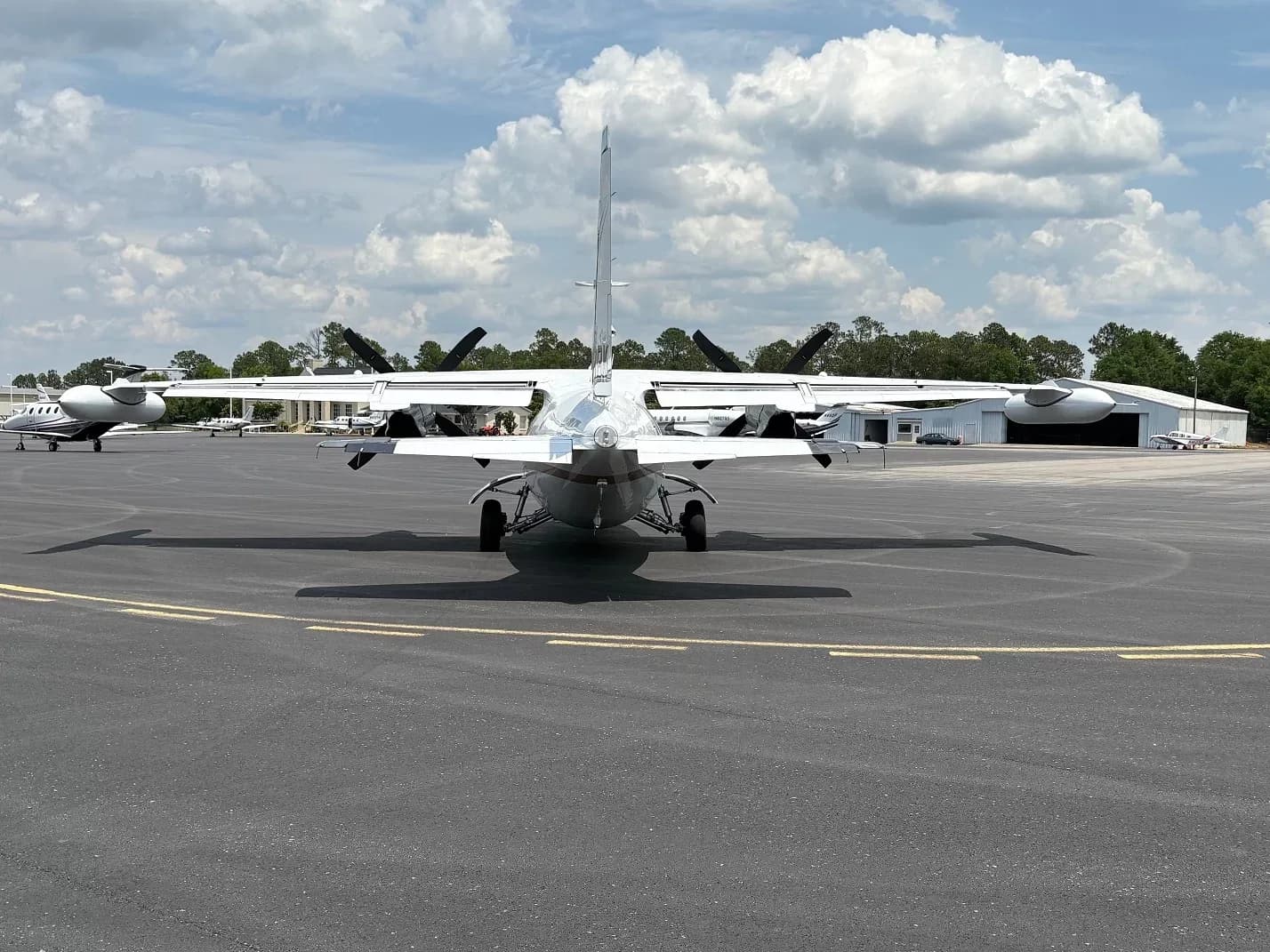 Rear view of a white twin-engine aircraft with four-blade propellers on an airfield under a cloudy sky.