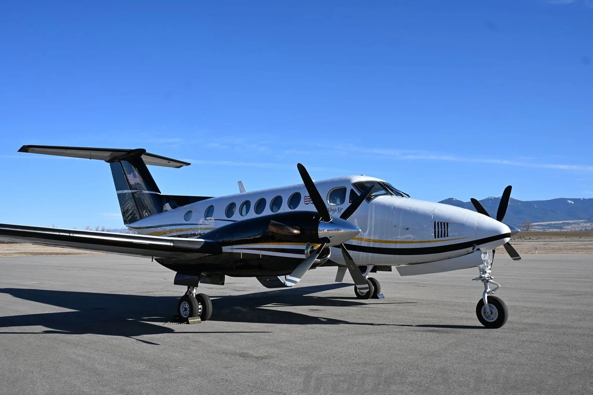 A white and black Beechcraft King Air 200 with gold stripes parked on an airfield, viewed from the front-left.