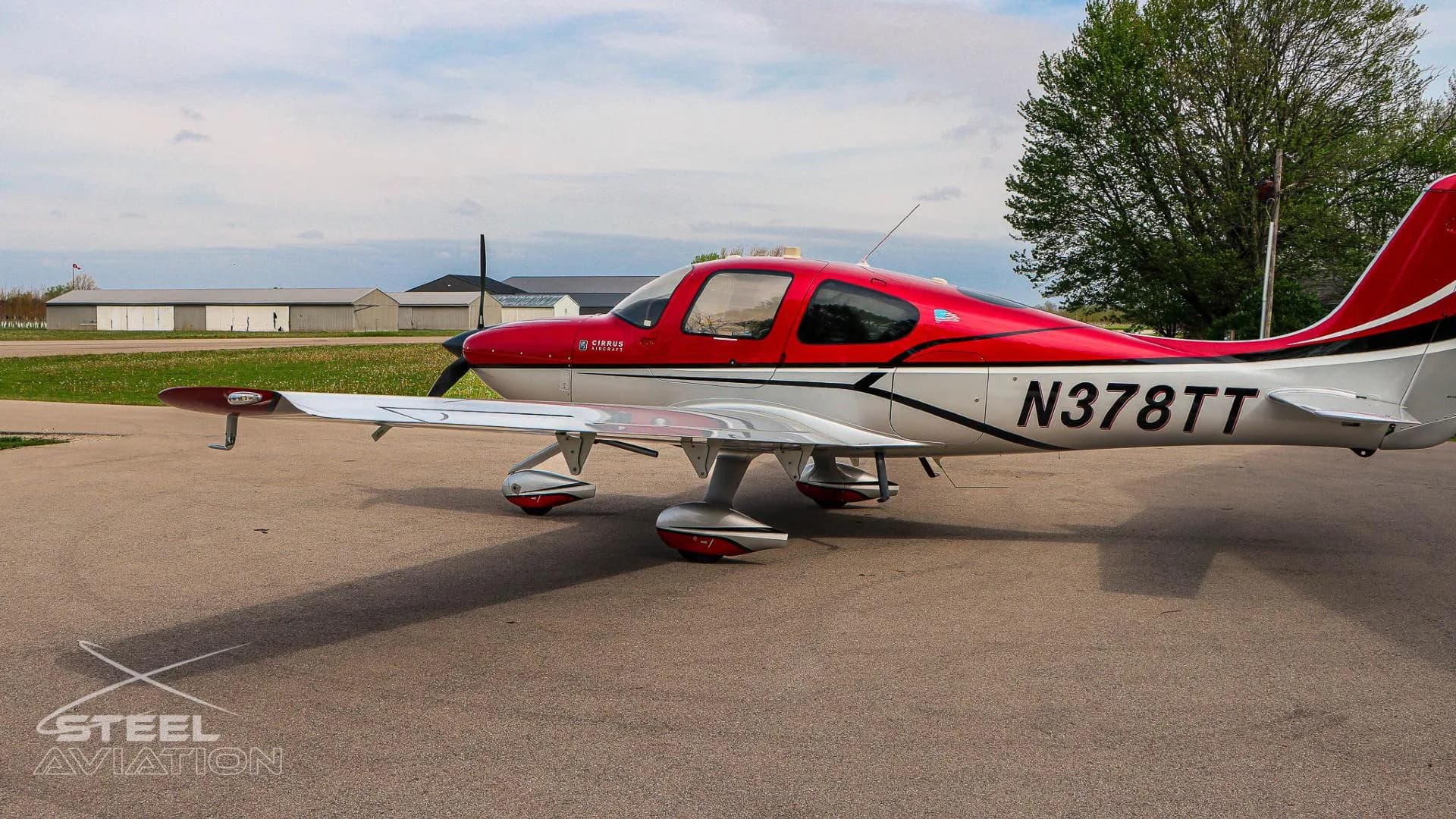 A red and white Cirrus SR22 aircraft with tail number N378TT is parked on an apron, viewed from the left side with other hangars in the distance.
