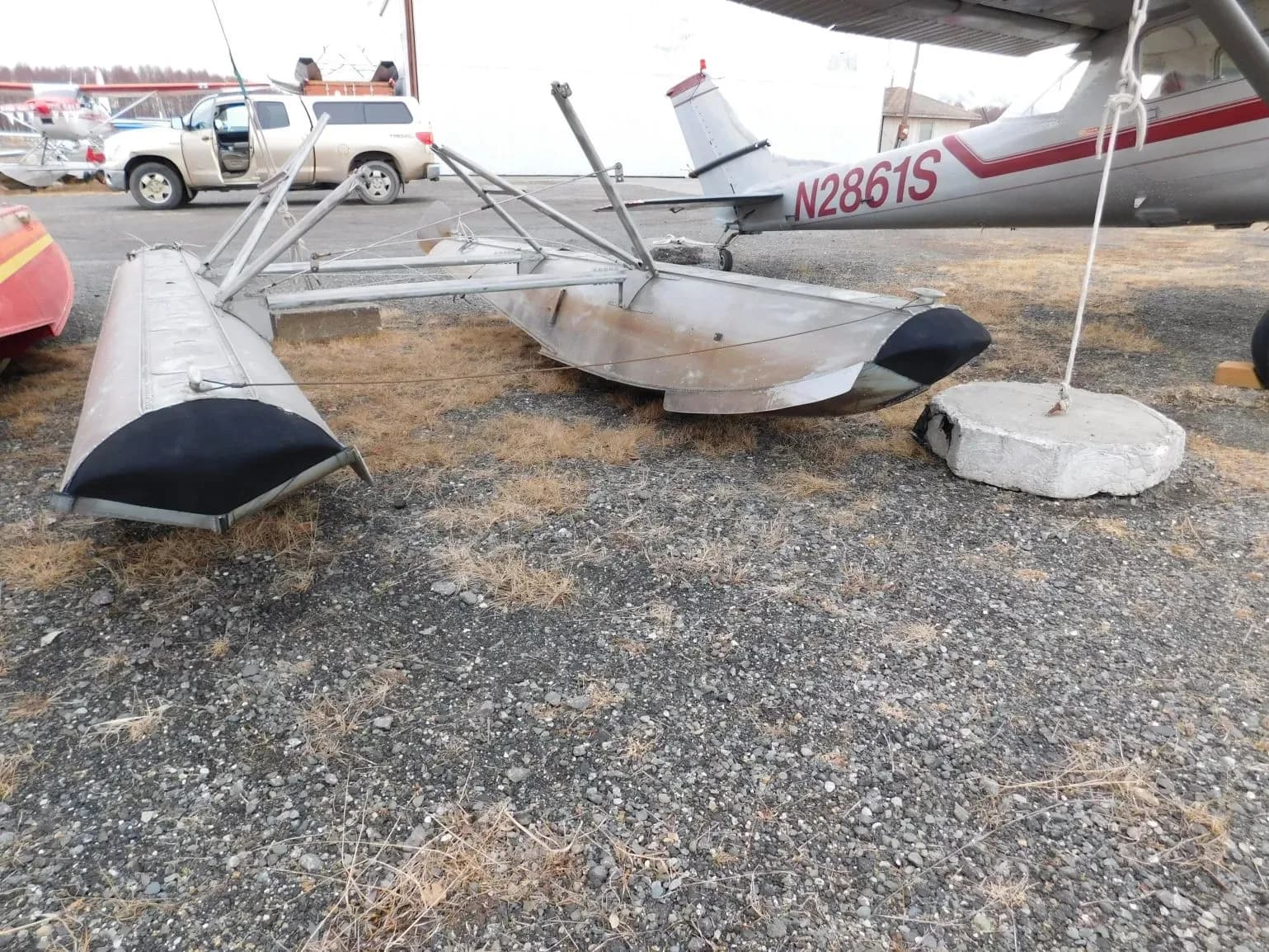 Two aircraft floats resting on gravel, with the tail of a silver and red aircraft (N2861S) visible in the background.