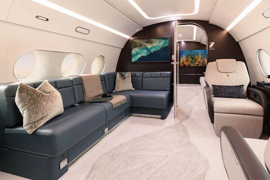 A private jet cabin with a large L-shaped blue leather sofa, decorative pillows, and two wall-mounted TV screens.