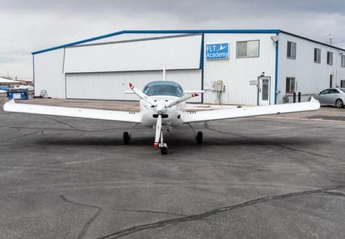 Front view of a white single-engine aircraft parked in front of a hangar.