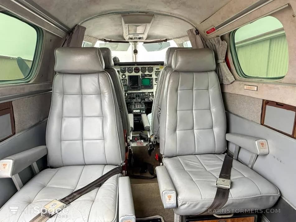 The interior cabin of an aircraft, showing two grey leather passenger seats with seatbelts, looking forward towards the cockpit.