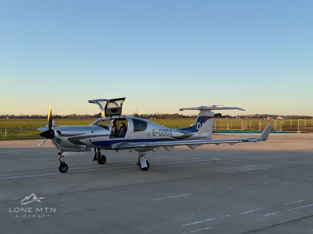 A silver and blue Diamond DA50 aircraft with registration G-DOGA and its gull-wing doors open, parked on an airfield.