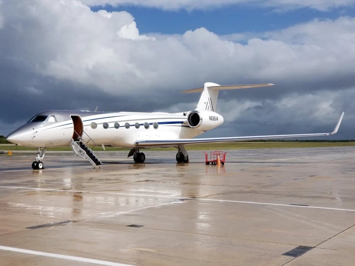 A white and silver Gulfstream G650 business jet with its door open and stairs extended, parked on a wet tarmac under a cloudy sky.