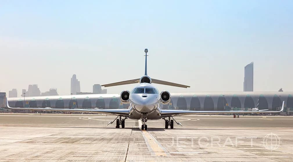 A white Dassault Falcon business jet with blue accents on the tail, seen from the front on a tarmac with a city skyline in the background.