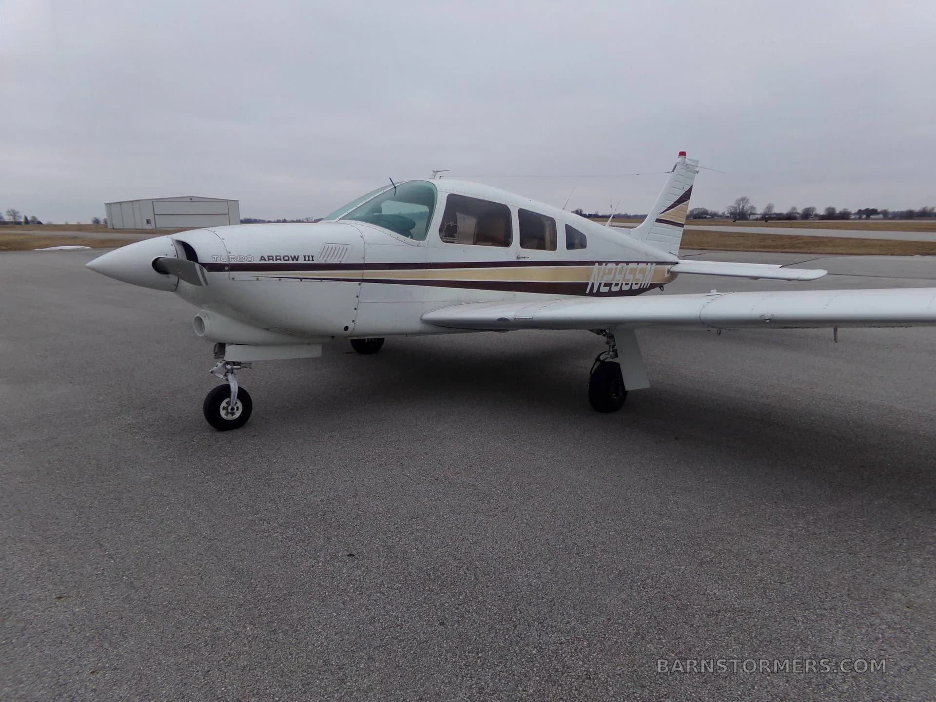 A white Piper Turbo Arrow III aircraft with brown and gold stripes, registration N2855M, is parked on an asphalt tarmac, viewed from the front-left.