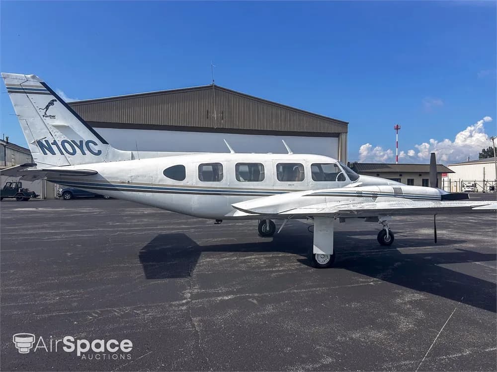 A white Piper Navajo aircraft with blue and gold stripes, registration N10YC, parked on an asphalt tarmac, viewed from the rear-left.