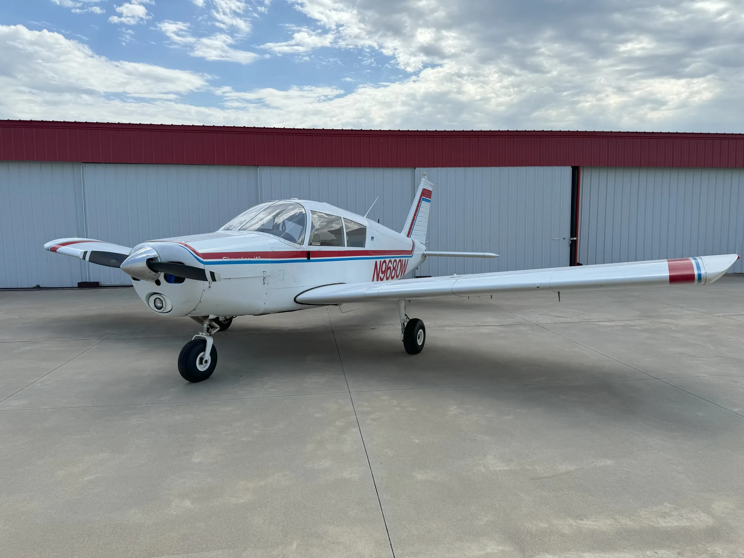 A white Piper Cherokee 140 aircraft with red and blue stripes is parked on a concrete tarmac in front of a hangar under a cloudy sky.